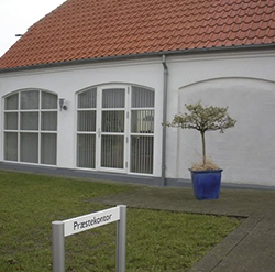 Weißes modernes Gemeindehaus mit großen Rundbogenfenstern und einem kleinen Topfbaum davor.