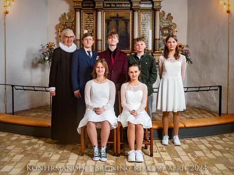 En gruppe unge konfirmander i hvide kjoler og jakkesæt poserer i en kirke.