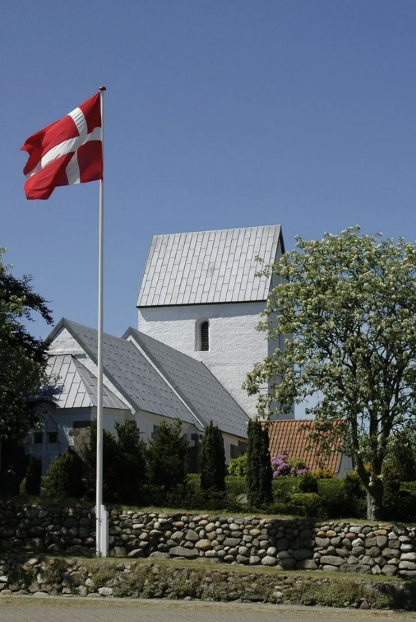 Hvid kirke bag en flagstang med det danske flag, Dannebrog, der vajer i vinden.