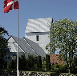 Weiße Kirche mit grauem Bleidach neben einer wehenden dänischen Flagge.