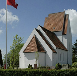 Weiße Dorfkirche mit rotem Ziegeldach neben einem hohen Fahnenmast unter blauem Himmel.