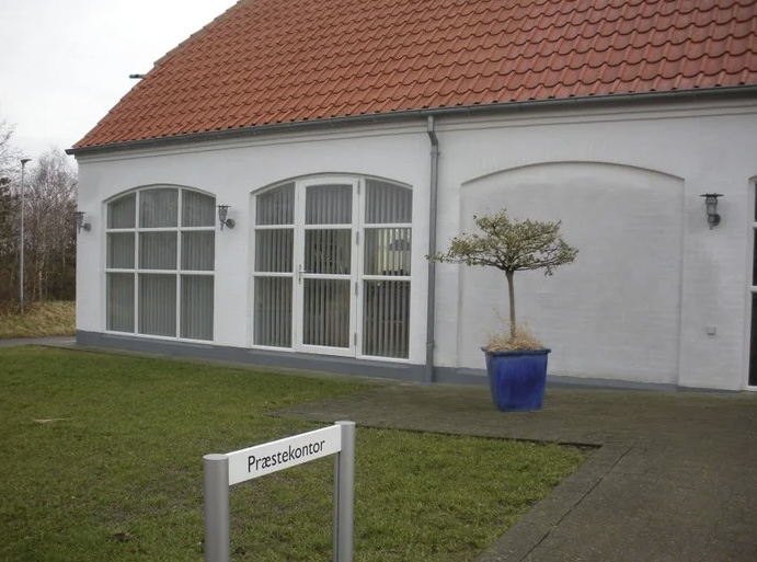 En hvid bygning med rødt tegltag, store sprossede vinduer og et mindre træ i en blå krukke.
