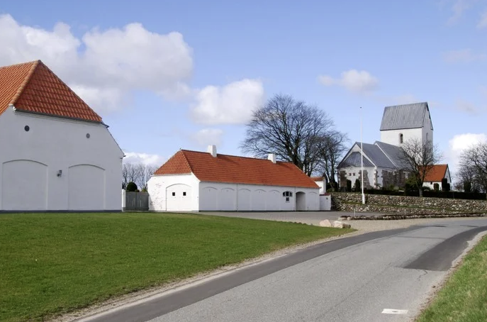 Vej forbi hvide bygninger med røde tage mod en landsbykirke.