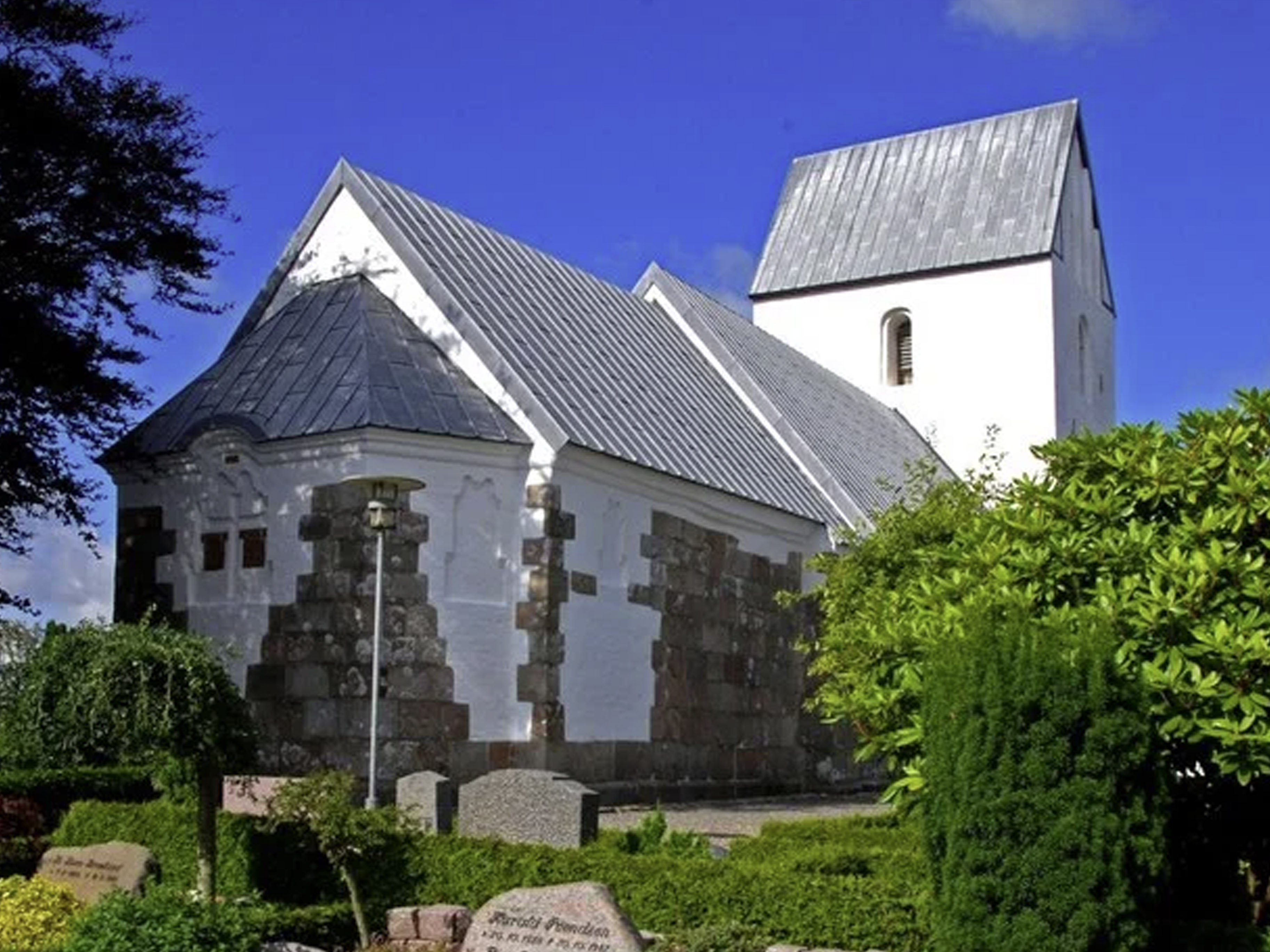 Hvid landsbykirke med metaltag og kampestensdetaljer under en blå himmel.