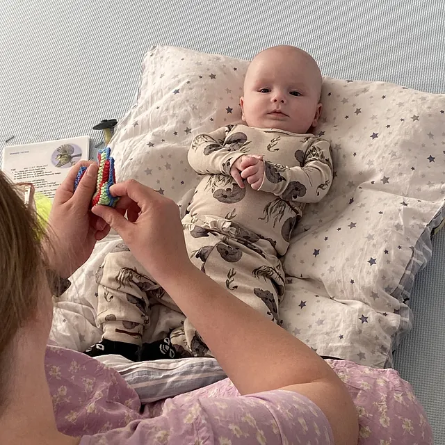 Baby liegt auf einem Kissen und schaut jemanden an, der ein Spielzeug hält.